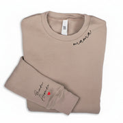 Folded beige sweatshirt with the word 'mama' written on the neckline and names on the right sleeve.